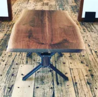 A Live Edge Tops wooden table with a natural edge design rests on a rustic wooden floor. The table showcases visible wood grain and is supported by metal legs, while the weathered look of the floor enhances its natural aesthetic.