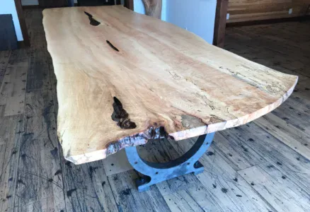 A large Live Edge Tops wooden table with a natural finish stands on a uniquely shaped metal base, featuring natural grain patterns and a few dark crevices. The floor and walls are crafted from rustic wood planks.