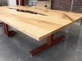 A large wooden table with a natural finish sits on red metal legs in a modern room with a concrete floor and brick walls. The tabletop features unique grain patterns and a central split design.