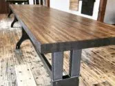 A long, rectangular wooden table with sturdy metal legs stands on a rustic hardwood floor. The tables smooth surface showcases the wood grain, contrasting with the industrial-style legs underneath. Another table is visible in the background.