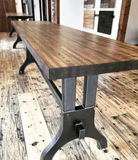 A long, rectangular wooden table with sturdy metal legs stands on a rustic hardwood floor. The tables smooth surface showcases the wood grain, contrasting with the industrial-style legs underneath. Another table is visible in the background.