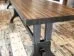 A long, rectangular wooden table with sturdy metal legs stands on a rustic hardwood floor. The tables smooth surface showcases the wood grain, contrasting with the industrial-style legs underneath. Another table is visible in the background.