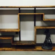 A wooden, geometrically designed bookcase with staggered shelves stands against a white wall. A small black statue of a cowboy is placed on the bottom right shelf, adding a decorative touch to the minimalist furniture piece.