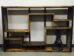 A wooden, geometrically designed bookcase with staggered shelves stands against a white wall. A small black statue of a cowboy is placed on the bottom right shelf, adding a decorative touch to the minimalist furniture piece.