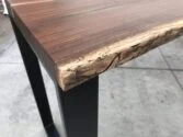 Close-up of a wooden table corner made from dark-stained wood with a natural live edge design, showcasing textured wood grain and unique markings. The table stands on black metal legs, set against a concrete floor background.