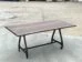A wooden table with a smooth, rectangular top and natural edges stands on a metal frame with four wheels, placed on a concrete surface. The background is an open outdoor area.