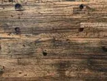 A close-up of a textured wooden surface with visible knots and grain patterns, displaying shades of brown and dark streaks, suggesting an aged or reclaimed wood look.