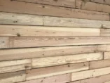 Wooden plank wall made of various shades of brown wood with visible grain patterns. The planks are arranged horizontally, creating a rustic texture and appearance. Some planks have knots and natural imperfections.