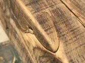 Close-up of a piece of aged, weathered wood with visible grain and a prominent crack. The wood surface shows various textures and patterns, highlighted by natural lighting.