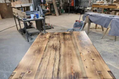 A handcrafted wooden table with a rustic finish stands in a workshop, surrounded by tools, workbenches, and equipment. The table features thick legs and a polished, textured tabletop with visible wood grain and knots.