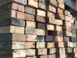 A stack of rough, weathered wooden beams piled high in a warehouse. The timber exhibits natural variations in color and age, with visible knots and grain patterns. The background shows part of a concrete wall.