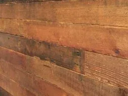 Close-up of a rustic wooden wall with staggered horizontal planks in various shades of brown. The surface shows natural grain and texture, with a small section of a red window frame visible at the top left corner.