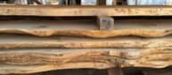 Stacked slabs of raw, unfinished wood with irregular, natural edges are neatly arranged. The wood appears to be in a workshop setting, hinting at carpentry or woodworking projects.