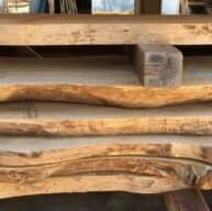 Stacked slabs of raw, unfinished wood with irregular, natural edges are neatly arranged. The wood appears to be in a workshop setting, hinting at carpentry or woodworking projects.