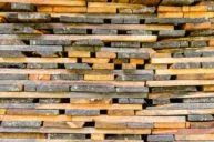 A close-up view of a neatly stacked pile of wooden planks, varying in color from light to dark brown. The planks are arranged horizontally, showing their textured surfaces and edges.
