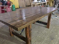 A large, rustic wooden table with thick legs and a patterned top sits in a workshop surrounded by tools, supplies, and industrial equipment.