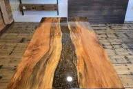 A polished wooden table with a black resin river running through the center stands on a wooden floor, with samples of wood panels displayed on the wall behind it.