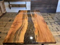 A polished wooden table with a black resin river running through the center stands on a wooden floor, with samples of wood panels displayed on the wall behind it.