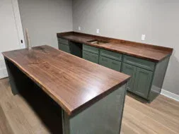 A kitchen with green cabinets and smooth, dark wooden countertops. An island with matching wood top and green base sits in the center. The room has light-colored walls and hardwood flooring.