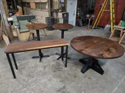 Four wooden tables with black metal legs—a round coffee table, a rectangular bench, and two small round side tables—are arranged in a workshop with tools, ladders, and industrial equipment in the background.