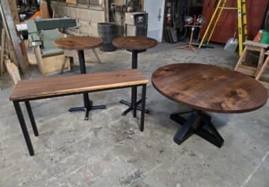 Four wooden tables with black metal legs—a round coffee table, a rectangular bench, and two small round side tables—are arranged in a workshop with tools, ladders, and industrial equipment in the background.