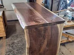 A polished wooden bar with a natural edge design is shown in a workshop setting. The wood has a rich, dark grain and is supported by a sturdy base. Workshop tools and materials are visible in the background.