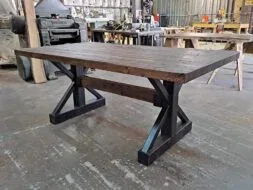 A sturdy wooden table with a thick, dark wood top and black metal X-shaped legs stands in a workshop setting. Various tools and equipment are visible in the background.
