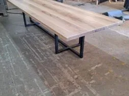 A long wooden table with a natural finish and black metal legs is set in a workshop. The surrounding space has woodworking tools and materials, visible in the background.
