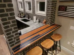 Home bar area with a brick wall, marble countertop, and three wooden stools. The bar top features an inlaid design with wood and dark stripes. Shelves with bottles and decor are visible behind the counter. Three small windows are set in the brick wall.