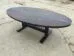 A large, oval wooden table with a dark finish and a cross pattern on the tabletop, situated on a rough, cracked concrete surface. The table has a sturdy black pedestal base with two wide legs.