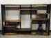 A wooden, geometrically designed bookcase with staggered shelves stands against a white wall. A small black statue of a cowboy is placed on the bottom right shelf, adding a decorative touch to the minimalist furniture piece.