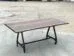A wooden table with a smooth, rectangular top and natural edges stands on a metal frame with four wheels, placed on a concrete surface. The background is an open outdoor area.