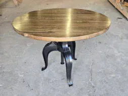 A round wooden table with a dark polished top and black metal legs stands on a concrete floor in a woodworking shop with tools and wooden planks in the background.